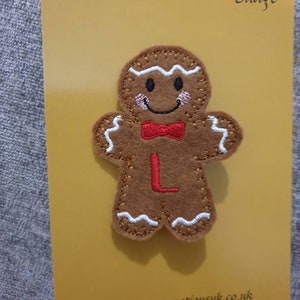 May include: A brown felt gingerbread man badge with white stitching, a red bow tie, and the letter "L". The badge is on a yellow card with the words "Patch" and "Badge". The website address www.manorcreationsuk.co.uk is visible.