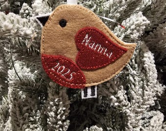 May include: A felt bird Christmas ornament with a tan body and red glitter wings. The ornament has the word "Nanna" on one wing and "2025" on the other. A white ribbon is attached for hanging.
