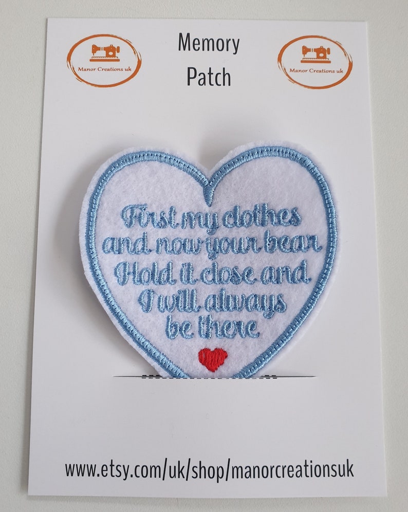 Op de afbeelding: Een hartvormige patch van wit vilt met een blauwe geborduurde rand. De patch heeft de tekst "First my clothes and now your bear Hold it close and I will always be there" in blauwe borduurwerk. Een klein rood vilt hart is in het midden onderaan de patch genaaid.