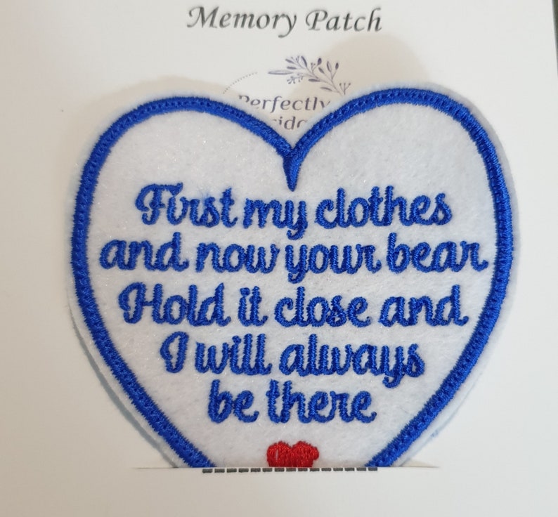 Op de afbeelding: Witte hartvormige viltpatch met blauwe geborduurde rand. De patch heeft de tekst "First my clothes and now your bear Hold it close and I will always be there" in blauwe borduurwerk. Een klein rood hart is onderaan de patch geborduurd.