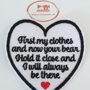 May include: White heart-shaped embroidered patch with black stitching and a red heart in the centre. The text reads "First my clothes and now your bear Hold it close and I will always be there."
