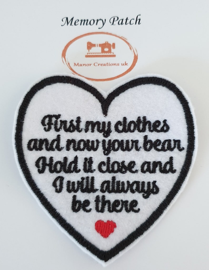 Op de afbeelding: Witte, hartvormige geborduurde patch met zwarte stiksels en een rood hart in het midden. De tekst luidt "First my clothes and now your bear Hold it close and I will always be there."