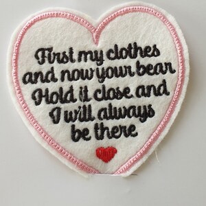 Op de afbeelding: Witte vilt hartvormige patch met roze stiksels rond de randen. De patch heeft een zwarte geborduurde tekst die luidt "First my clothes and now your bear Hold it close and I will always be there". Een klein rood hart is geborduurd aan de onderkant van de patch.