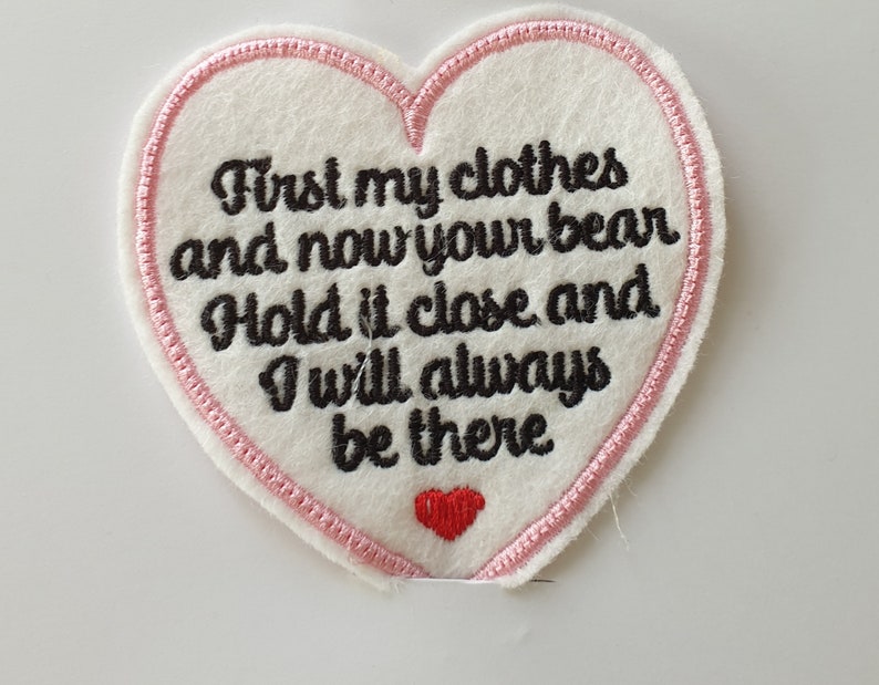Op de afbeelding: Witte vilt hartvormige patch met roze stiksels rond de randen. De patch heeft een zwarte geborduurde tekst die luidt "First my clothes and now your bear Hold it close and I will always be there". Een klein rood hart is geborduurd aan de onderkant van de patch.