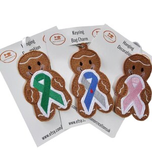 May include: Three gingerbread man fabric charms with ribbon designs. One has a green ribbon, one a blue and grey ribbon, and one a pink ribbon. Each charm is attached to a white card with text and a metal ring for hanging.