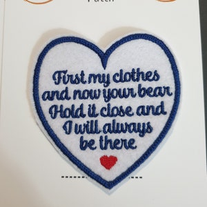 Op de afbeelding: Witte vilt hartvormige patch met blauwe geborduurde rand. De patch heeft de tekst "First my clothes and now your bear Hold it close and I will always be there" in zwarte borduurwerk. Een klein rood hart is geborduurd in het midden van de patch.