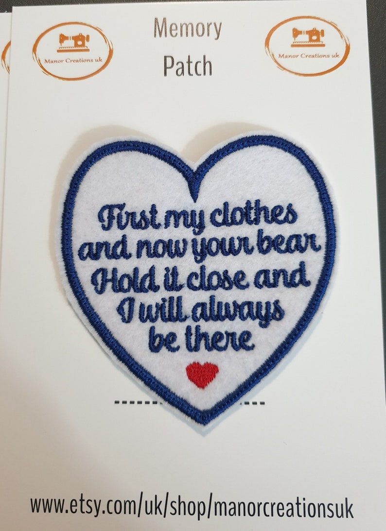 Op de afbeelding: Witte vilt hartvormige patch met blauwe geborduurde rand. De patch heeft de tekst "First my clothes and now your bear Hold it close and I will always be there" in zwarte borduurwerk. Een klein rood hart is geborduurd in het midden van de patch.