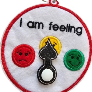 May include: A white felt circle with a red border and the text "I am feeling" in black. There are three embroidered faces: a red sad face, a yellow neutral face, and a green happy face. A black arrow with a white button in the centre can be moved to point to each face.