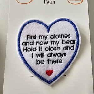 May include: A heart-shaped memory patch with a white felt base and a blue stitched border. The patch features embroidered text: "First my clothes and now my bear, Hold it close and I will always be there." A small red heart is at the bottom.