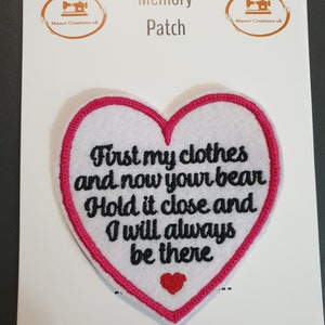 Op de afbeelding: Witte, hartvormige geborduurde patch met roze stiksels. De patch zegt "First my clothes and now your bear. Hold it close and I will always be there".