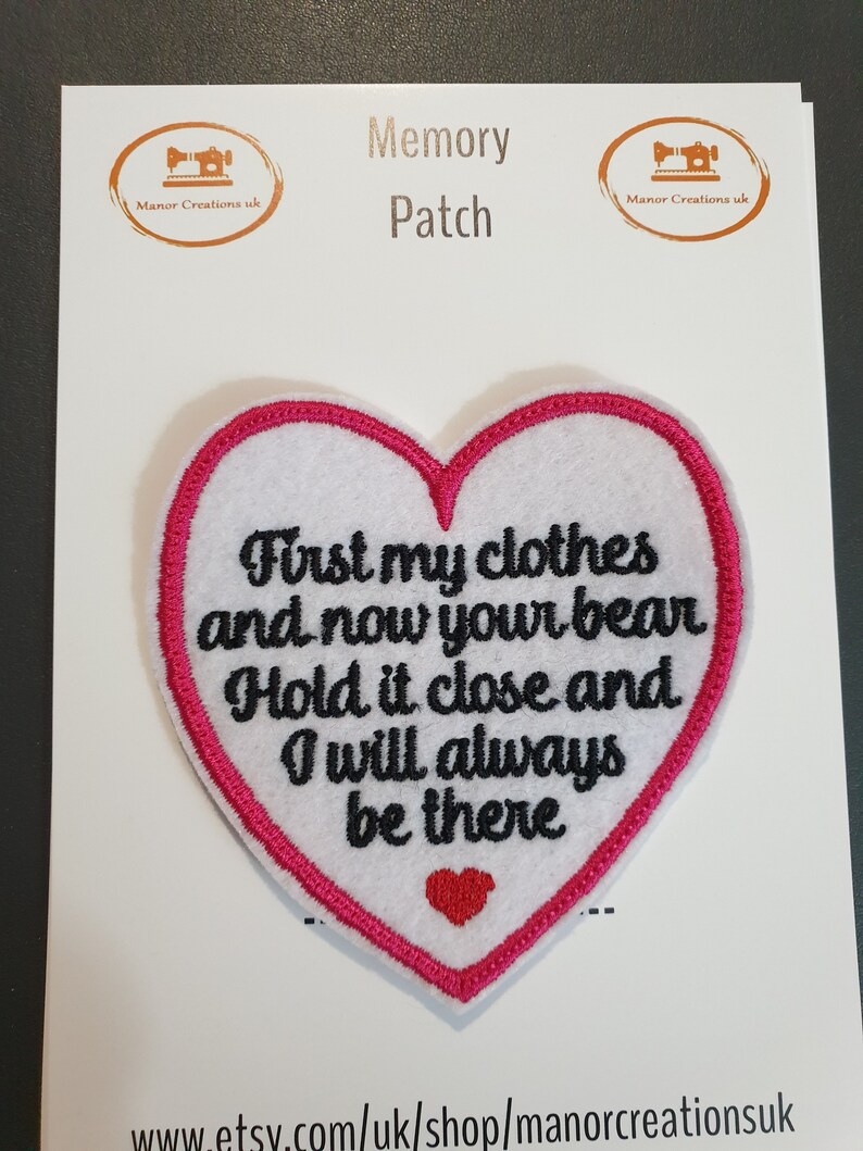 Op de afbeelding: Witte, hartvormige geborduurde patch met roze stiksels. De patch zegt "First my clothes and now your bear. Hold it close and I will always be there".