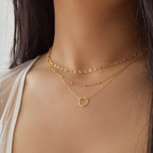 May include: A gold layered necklace with a delicate circle pendant. The necklace is worn by a person with long dark hair.