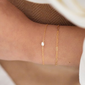 May include: A delicate gold chain bracelet with a single pearl charm. The bracelet is layered with a gold chain link bracelet.
