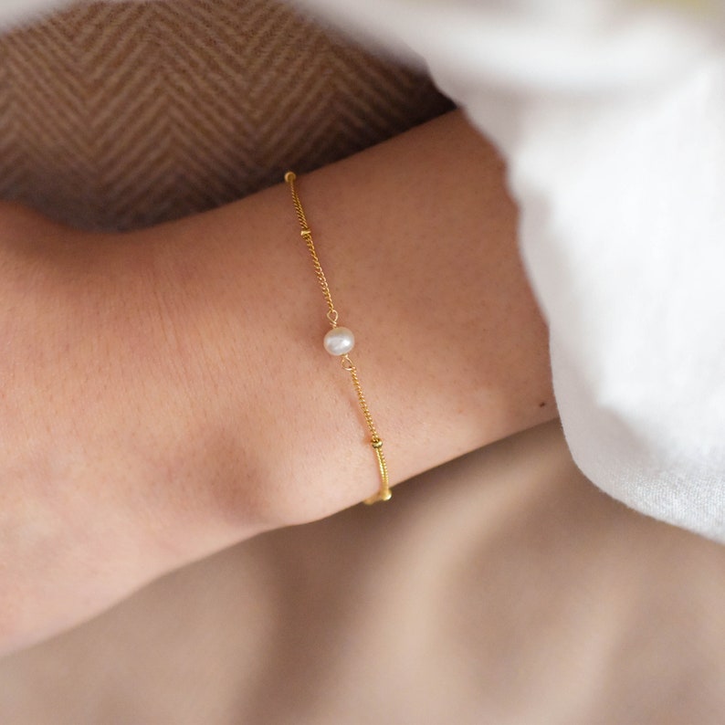 Pode incluir: Um delicado bracelete de corrente dourada com um &uacute;nico charme de p&eacute;rola branca. O bracelete &eacute; simples e elegante, perfeito para o uso di&aacute;rio.