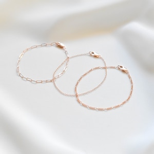 Puede incluir: Dos delicados brazaletes de cadena de oro, uno con una cadena simple y el otro con un dise&ntilde;o m&aacute;s intrincado, se muestran sobre un fondo blanco.