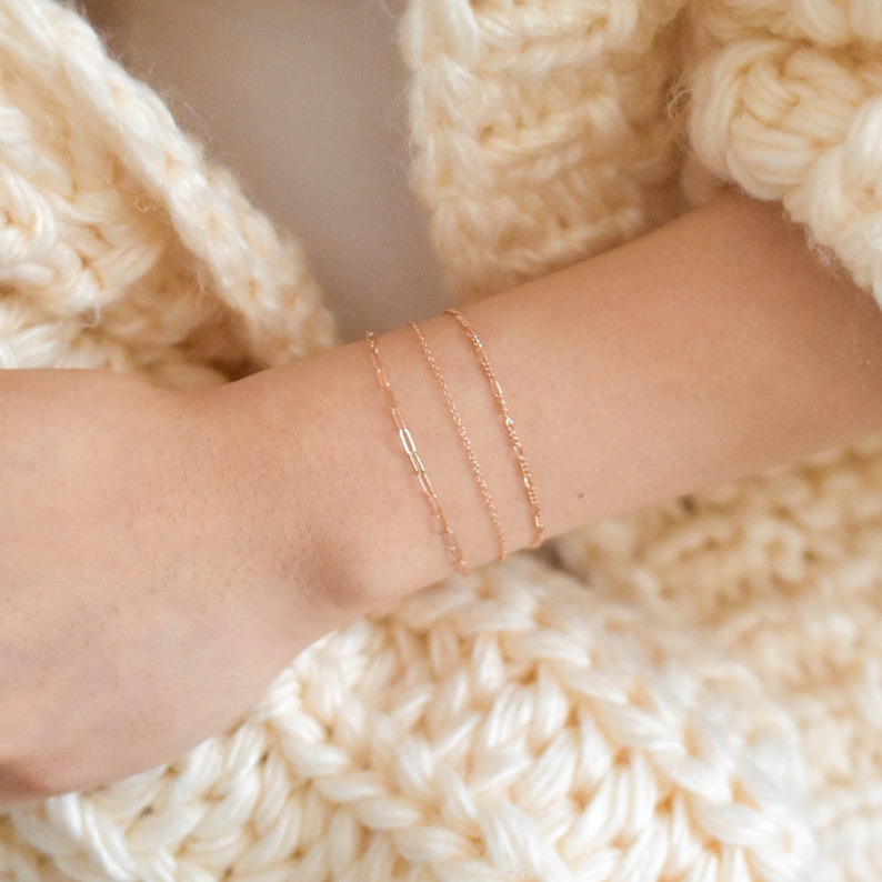 Puede incluir: Tres pulseras de cadena doradas apiladas en la mu&ntilde;eca de una persona. Las pulseras son delicadas y tienen un dise&ntilde;o simple.