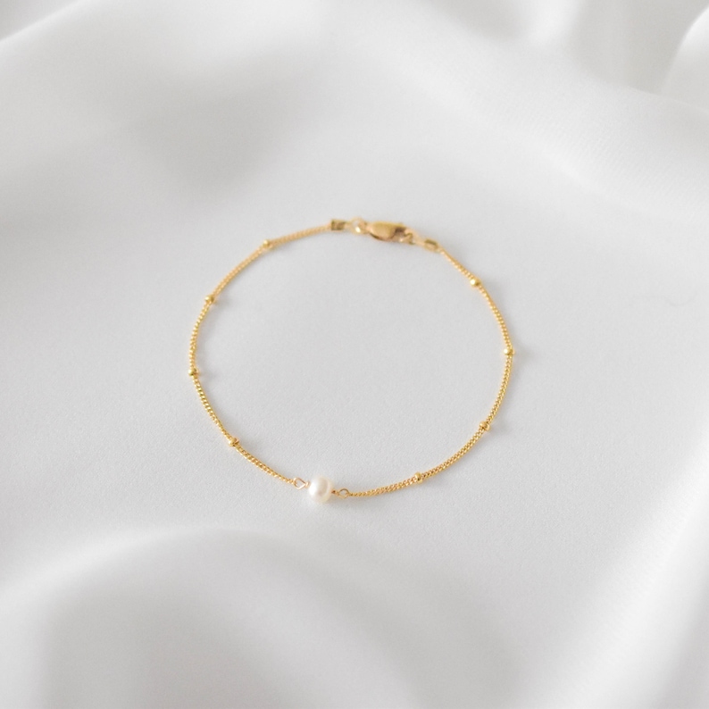 Pode incluir: Um delicado bracelete de corrente dourada com um &uacute;nico charme de p&eacute;rola. O bracelete &eacute; feito de pequenas contas redondas e tem um design simples e elegante.