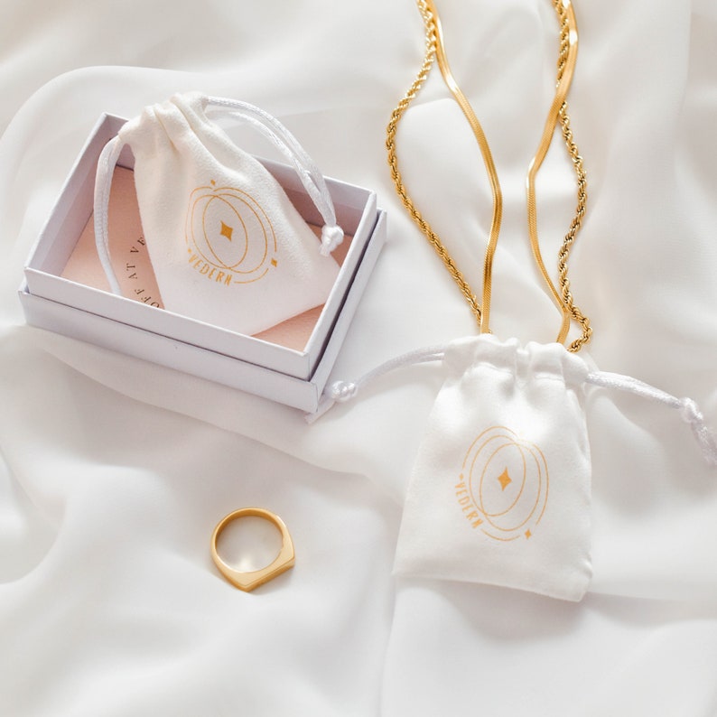 Pode incluir: Duas bolsas de veludo branco com letras douradas que dizem "VEDIRA" e um colar de corrente dourada. As bolsas est&atilde;o sobre uma superf&iacute;cie branca.