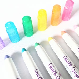May include: A collection of colorful chalk crayons with matching translucent caps. The crayons are arranged in a rainbow of colors, including purple, blue, green, yellow, orange, and pink. Each crayon has the words "Chalk Crayons LOVE DESIGNS" printed on the side.
