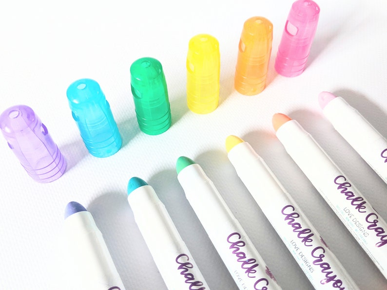 May include: A collection of colorful chalk crayons with matching translucent caps. The crayons are arranged in a rainbow of colors, including purple, blue, green, yellow, orange, and pink. Each crayon has the words "Chalk Crayons LOVE DESIGNS" printed on the side.