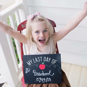 May include: A young girl with blonde hair and blue eyes sits in a red chair, holding a chalkboard sign that reads "MY LAST DAY OF Preschool 5/24/18." She is smiling with her arms raised in the air. The background includes a white porch railing.
