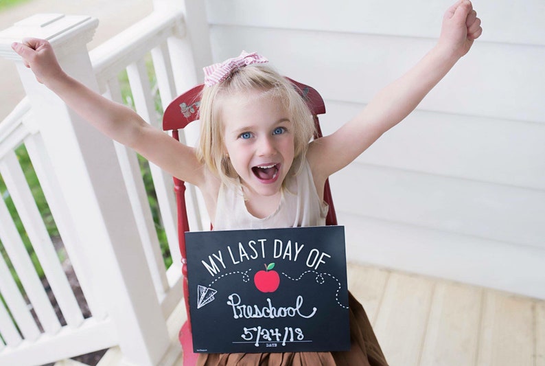 May include: A young girl with blonde hair and blue eyes sits in a red chair, holding a chalkboard sign that reads "MY LAST DAY OF Preschool 5/24/18." She is smiling with her arms raised in the air. The background includes a white porch railing.