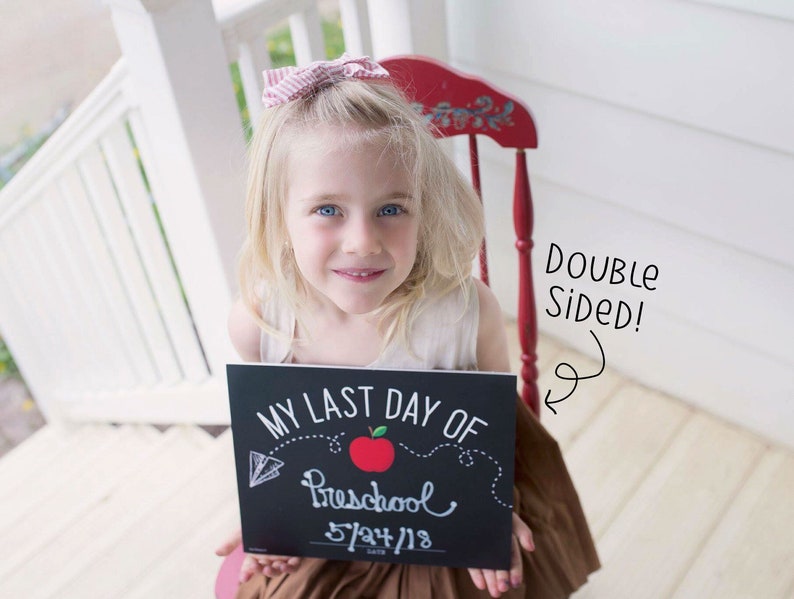 May include: A young girl with blonde hair and blue eyes smiles while holding a chalkboard sign. The sign reads "MY LAST DAY OF Preschool 5/24/18" with a red apple and a dotted line design. The sign is double-sided.