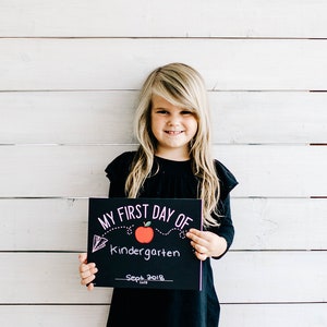 May include: A young girl in a black dress holds a black sign that reads "MY FIRST DAY OF Kindergarten" with a red apple illustration and the date "Sept 2018". The background is a white wooden wall.