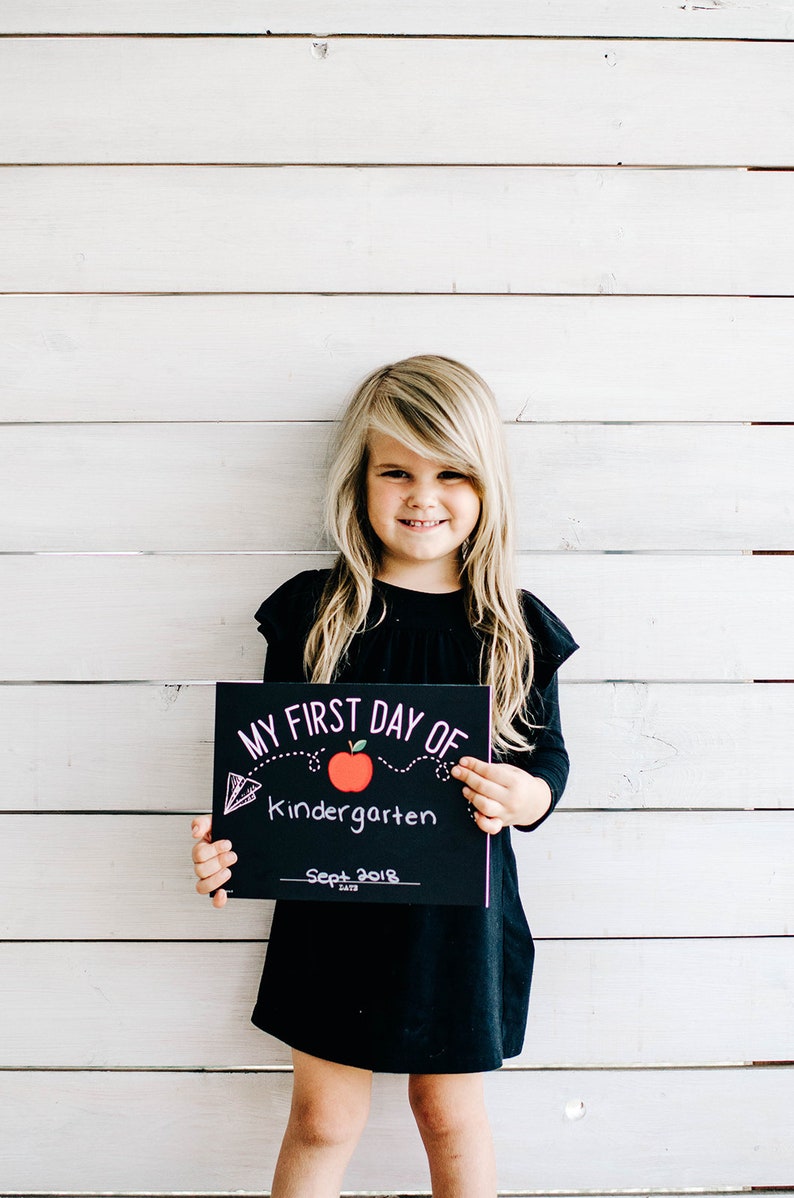 May include: A young girl in a black dress holds a black sign that reads "MY FIRST DAY OF Kindergarten" with a red apple illustration and the date "Sept 2018". The background is a white wooden wall.