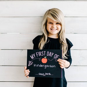 May include: A smiling child with long blonde hair holds a black chalkboard sign that reads "MY FIRST DAY OF Kindergarten Sept 2018" with a red apple graphic. The child wears a black long-sleeved shirt. The background is a white wooden wall.