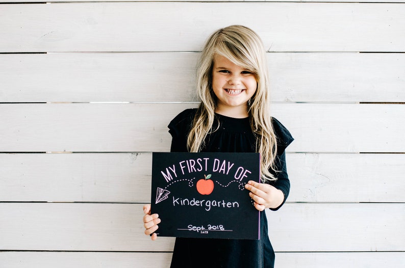 May include: A smiling child with long blonde hair holds a black chalkboard sign that reads "MY FIRST DAY OF Kindergarten Sept 2018" with a red apple graphic. The child wears a black long-sleeved shirt. The background is a white wooden wall.