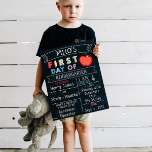 May include: A boy holds a chalkboard sign that says "Milo's First Day of Kindergarten". The sign has a red apple, the words "Kindergarten", "My School", "Henry James", "I am", "Strong + Powerful", "When I grow up I want to be Excavator Operator", "I am 4 years old", "I love Playing with Dinosaurs Cars My Stuffy", and "Sept 6, 2019".