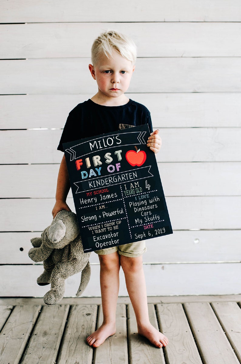 May include: A boy holds a chalkboard sign that says "Milo's First Day of Kindergarten". The sign has a red apple, the words "Kindergarten", "My School", "Henry James", "I am", "Strong + Powerful", "When I grow up I want to be Excavator Operator", "I am 4 years old", "I love Playing with Dinosaurs Cars My Stuffy", and "Sept 6, 2019".