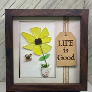 May include: A framed artwork featuring a yellow flower made of sea glass in a pot with a bee, and a brown tag with the text "LIFE is Good".