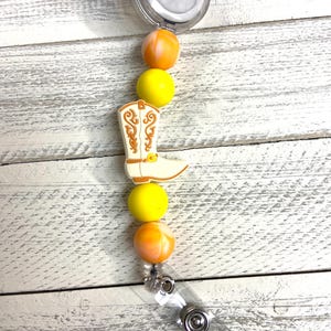 Puede incluir: Un carrete de insignia retráctil con cuentas amarillas y naranjas con un dije de bota blanca con detalles marrones.