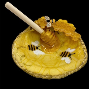 Puede incluir: Un plato de miel decorativo amarillo con borde dorado, con un cucharón de miel de madera. El plato está adornado con abejas pintadas y un diseño de panal. Una abeja enjoyada descansa sobre el panal. El plato tiene unos 13 cm de diámetro.