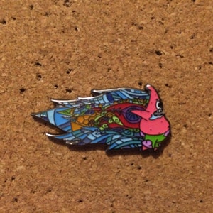 May include: A colorful enamel pin featuring a cartoon character with a pink body, blue eyes, and a red mouth. The character is surrounded by a blue, green, and red swirl pattern.