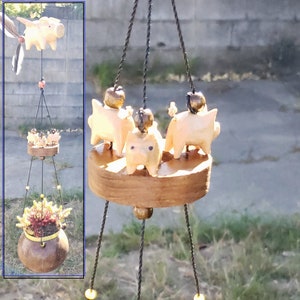 May include: A hanging planter with three wooden pig figurines on the top tier and a succulent plant in a coconut shell on the bottom tier. The planter is suspended by three black cords with yellow beads.