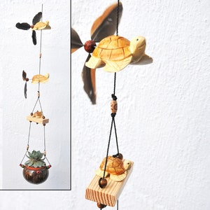 Peut inclure: Un carillon en bois en forme de tortue avec trois tortues et un pot en noix de coco avec une plante succulente. Les tortues sont peintes en brun et ont des yeux noirs. Le carillon est suspendu à une ficelle noire avec des perles en bois.