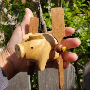May include: A wooden pig figurine with brown wings and a yellow bead on the tail. The pig is painted a light brown color and has black eyes.