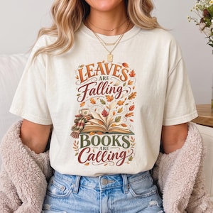 May include: An off-white short-sleeve t-shirt featuring a graphic print of an open book surrounded by colorful autumn leaves. The text reads LEAVES ARE FALLING BOOKS ARE CALLING. The design includes a small mug and a plaid scarf resting on the books.