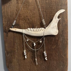 May include: A white animal jawbone suspended by silver-toned metal chains. The piece features clear faceted beads, small white crescent moon charms, and a clear crystal point pendant hanging from the lower chains, creating a layered design.