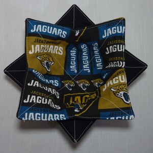 May include: A black and gold fabric pot holder with the Jacksonville Jaguars logo and the words "Jacksonville Jaguars" repeated throughout. The pot holder is folded into a star shape.