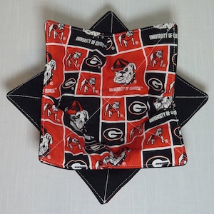 May include: A quilted bowl cozy featuring a red, black, and white University of Georgia Bulldogs pattern. The square-shaped cozy has a black star-shaped base and is designed to hold a bowl. The fabric displays the university's logo and mascot.