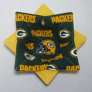 May include: A green and yellow quilted bowl cozy featuring the Green Bay Packers logo and text. The cozy is square with a star-shaped design, with the Packers logo and text in yellow on a green background.