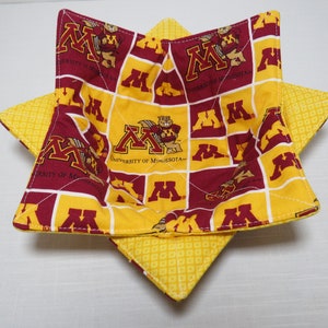 May include: A quilted fabric bowl cozy featuring a repeating pattern of the University of Minnesota logo and mascot in maroon and gold. The interior is divided into a grid of squares with the school branding, while the exterior is yellow fabric.