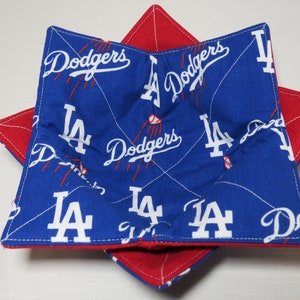 May include: Two fabric bowl cozies, one inside the other, in a star shape. The top cozy is blue with white "Dodgers" text and "LA" logos. The bottom cozy is red. Both are stitched with white thread.