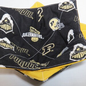 May include: A fabric bowl cozy featuring a black interior with a repeating Purdue Boilermakers logo, basketball, and football helmet print in gold and white. The exterior is solid yellow fabric with white cross-stitch quilting throughout the square shape.