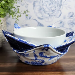 May include: A white ceramic bowl sits inside a blue and white floral patterned bowl cozy. The cozy has a dark blue interior and a light blue and white floral exterior. The background features a floral pattern in blue and white.