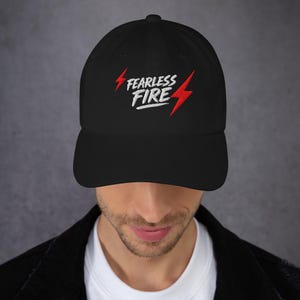 以下が含まれることがあります： 黒い野球帽。白い文字で「FEARLESS FIRE」と刺繍され、赤い稲妻のアクセントが付いています。帽子はカーブしたつばと、クラシックで調整可能なデザインです。
